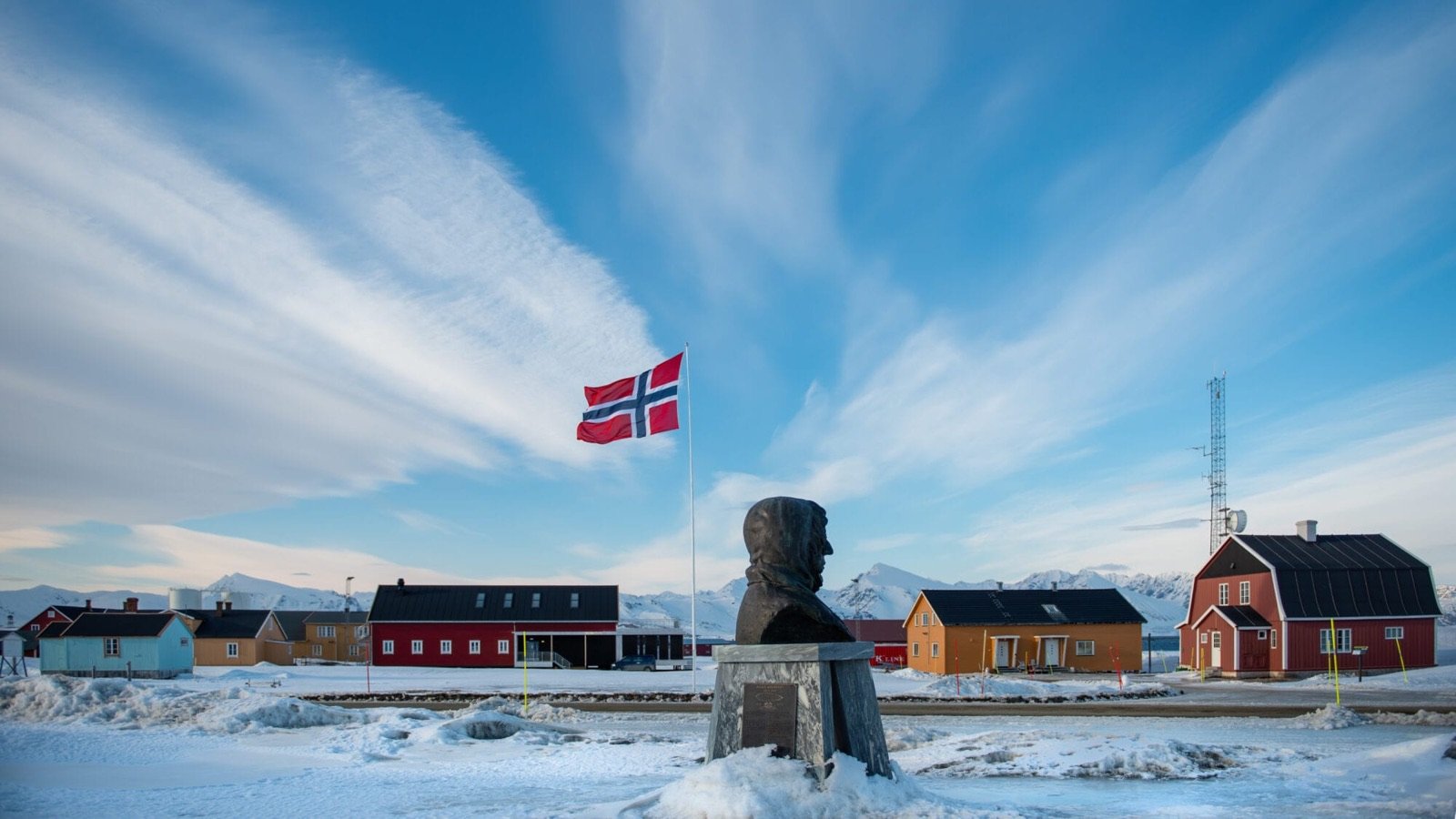 The red and yellow wooden houses of Ny-Ålesund in winter, with a Norwegian flag and a bust on a plinth